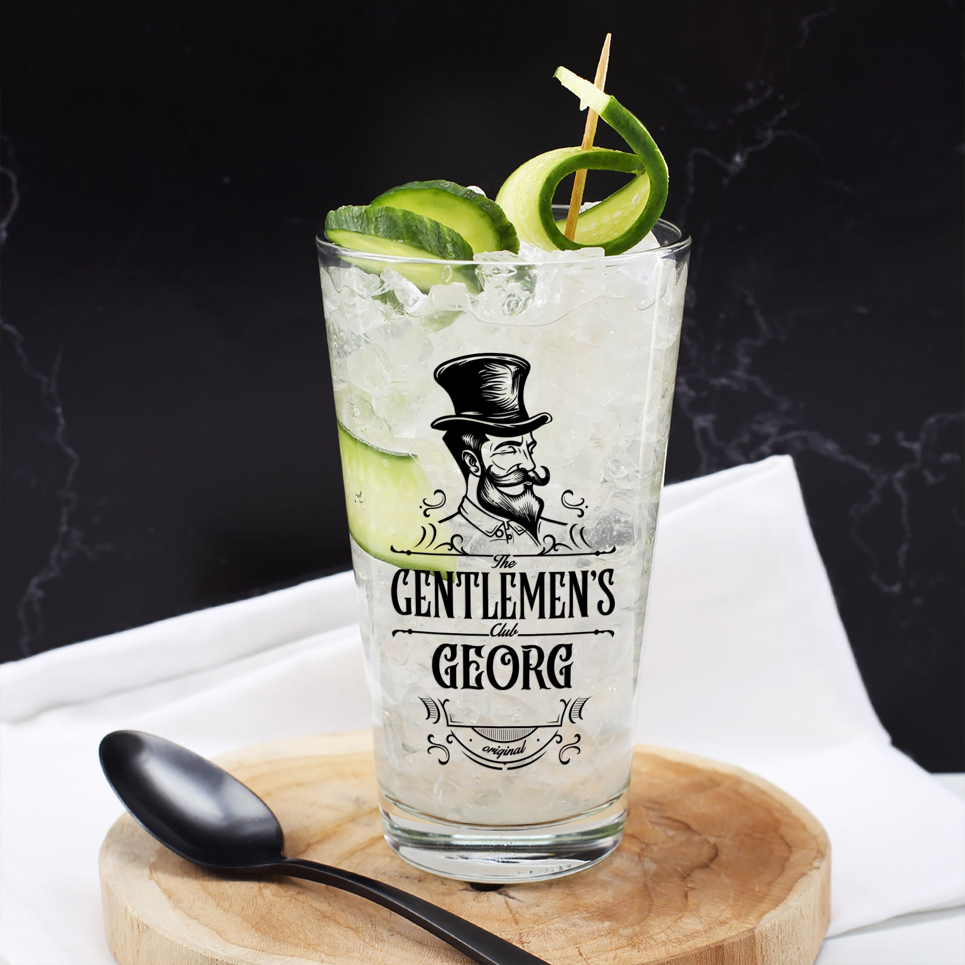 Trinkglas The Gentlemen’s Club mit Name Georg, befüllt mit Eis, Gurke und Drink, auf Holzteller mit schwarzem Löffel