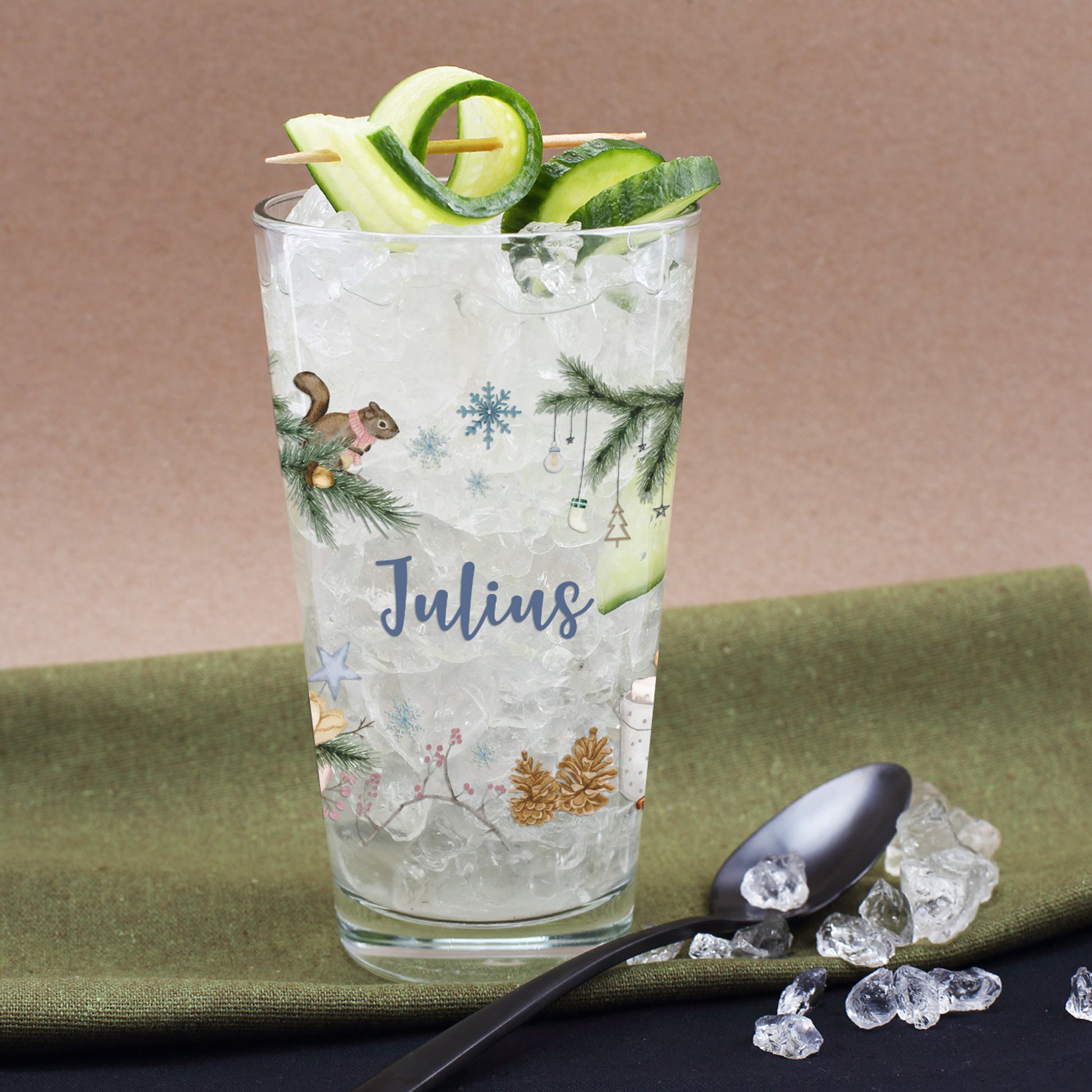 Nahaufnahme eines personalisierten Glases mit Tannenzweigen und Eichhörnchen-Motiv, dekoriert mit Gurken und Eiswürfeln