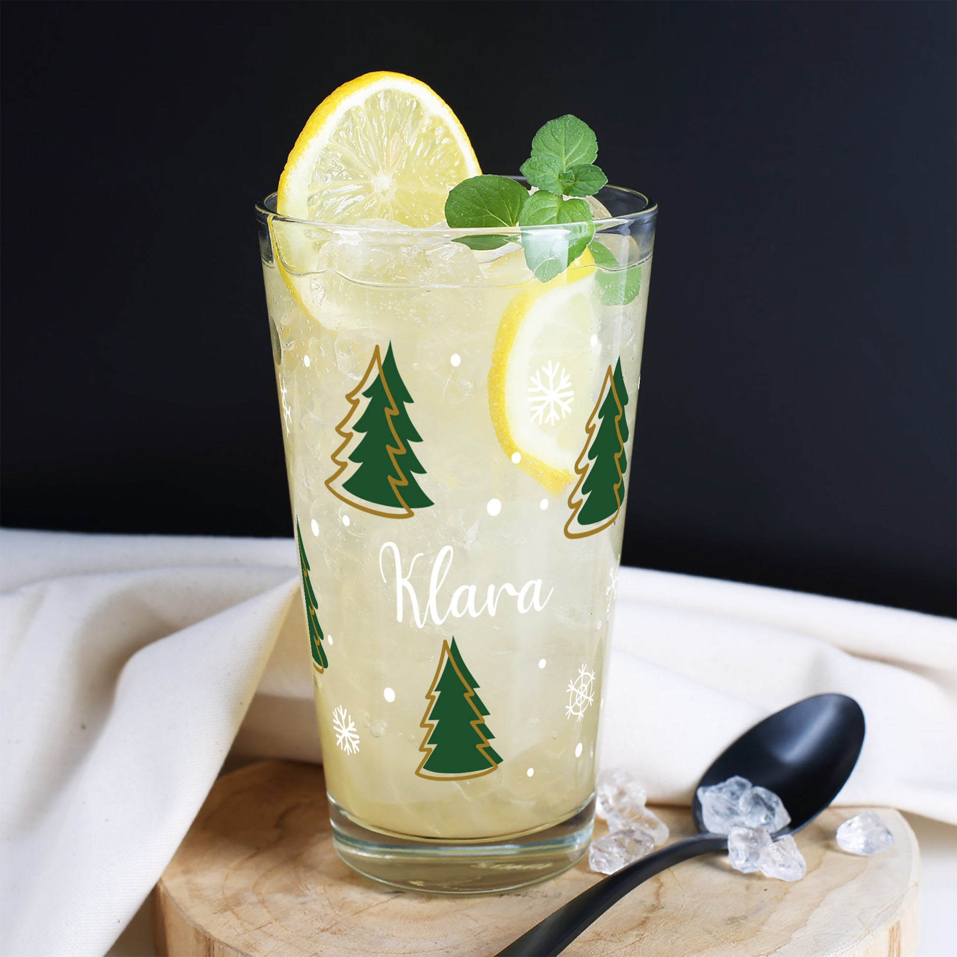 Personalisierbares Weihnachtsglas mit Tannenbäumen, Schneeflocken und Namen – auf rustikaler Holzscheibe arrangiert.