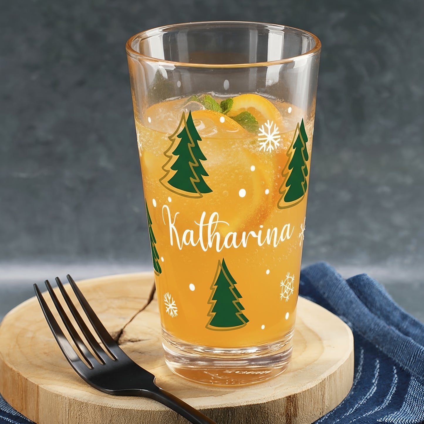 Weihnachtliches Trinkglas mit grünem Tannenbaum-Design und weißen Schneeflocken, personalisiert mit dem Namen Katharina, befüllt mit orangem Getränk, Minze und Orangenscheiben.