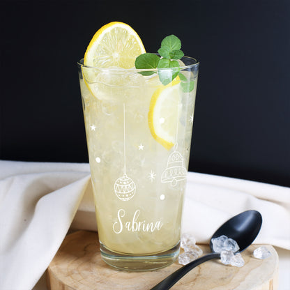 Personalisierbares Trinkglas mit weißen Weihnachtsmotiven – Glocke, Kugeln und Sternen – gefüllt mit Zitronengetränk und Eis, dekoriert mit Zitronenscheibe und Minze.