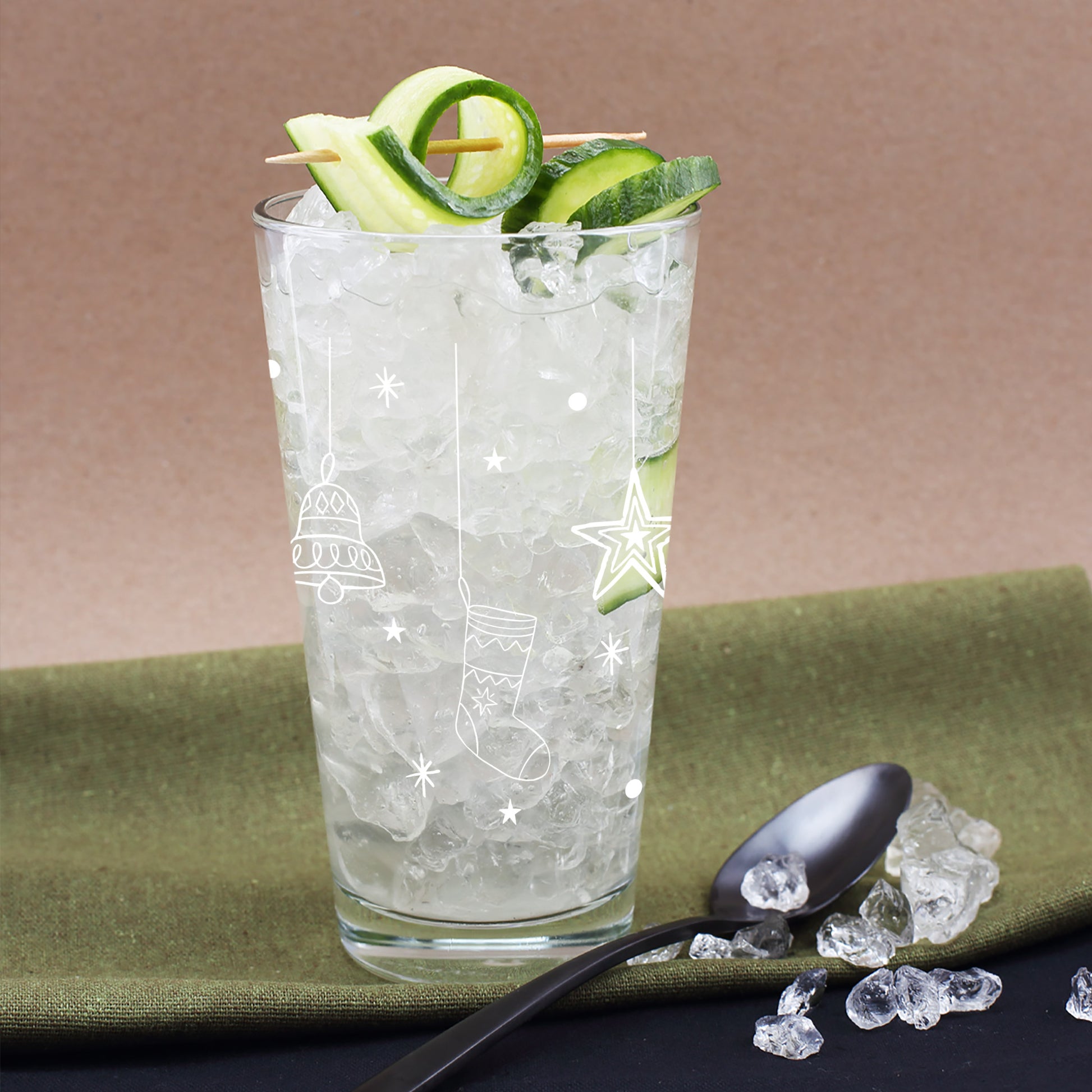 Weihnachtliches Glas mit weißen Glocken-, Stern- und Strumpfmotiven, gefüllt mit Eis und Gurkendekoration, festlich arrangiert auf grünem Untergrund.