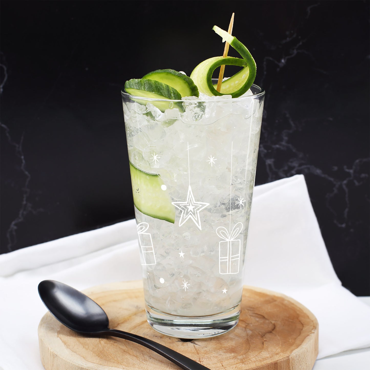 Personalisiertes Trinkglas mit weißen Weihnachtsmotiven – Sternen, Geschenken und Eiskristallen – befüllt mit Eiswürfeln und Gurke.