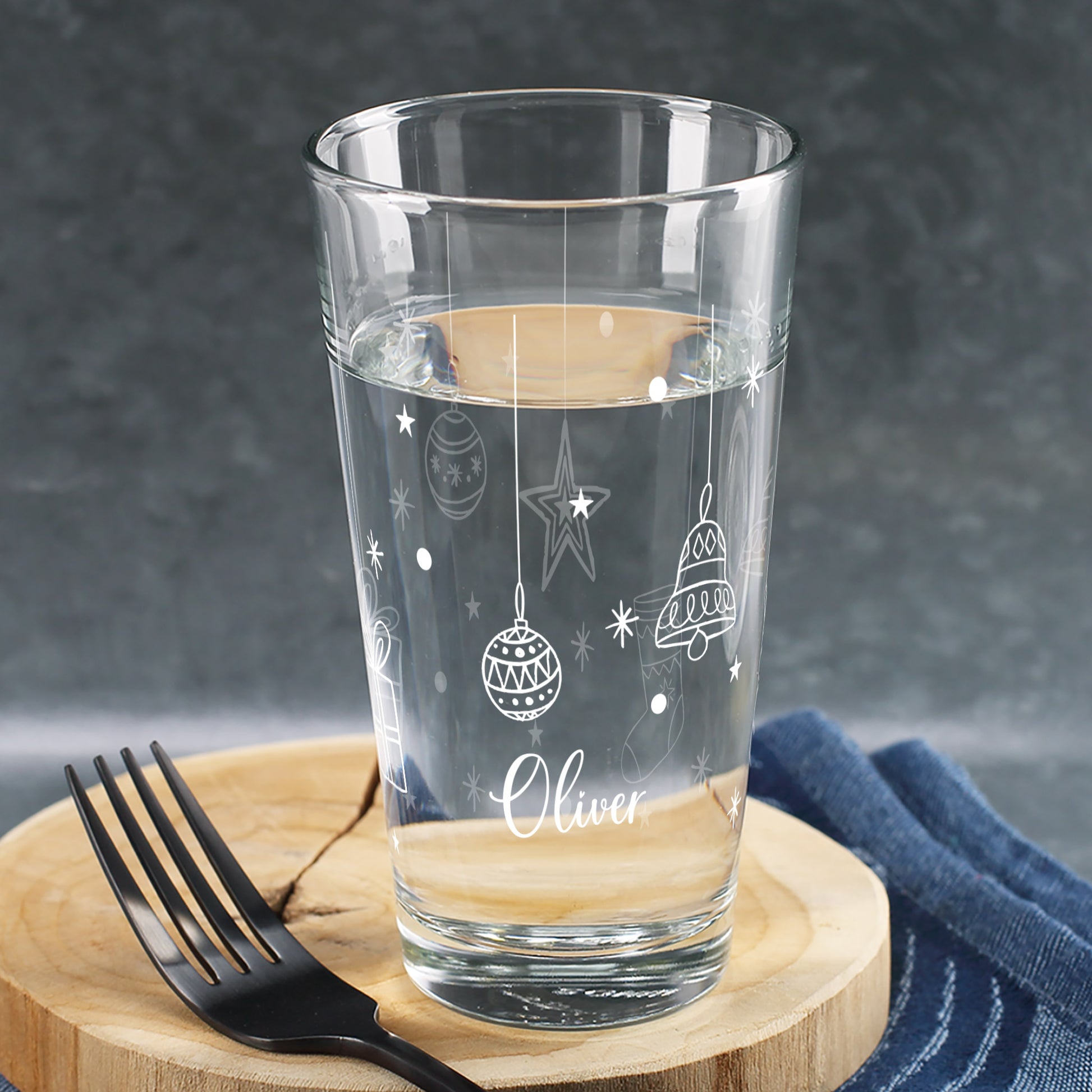 Personalisierbares Weihnachtsglas mit weißen Kugel-, Stern- und Glockenmotiven, dekoriert mit Wasserfüllung und Namen, auf rustikalem Holzuntergrund.