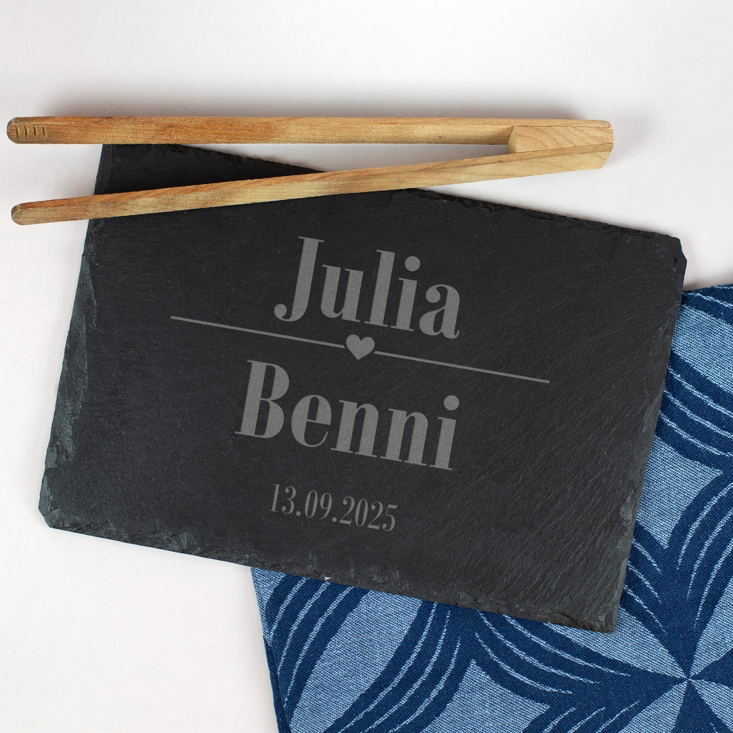 Schieferplatte mit Gravur „Julia & Benni“ und Datum 13.09.2025, verziert mit Herzsymbol, auf blauem Stofftuch und mit Grillzange dekoriert