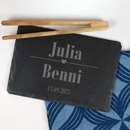 Schieferplatte mit Gravur „Julia & Benni“ und Datum 13.09.2025, verziert mit Herzsymbol, auf blauem Stofftuch und mit Grillzange dekoriert