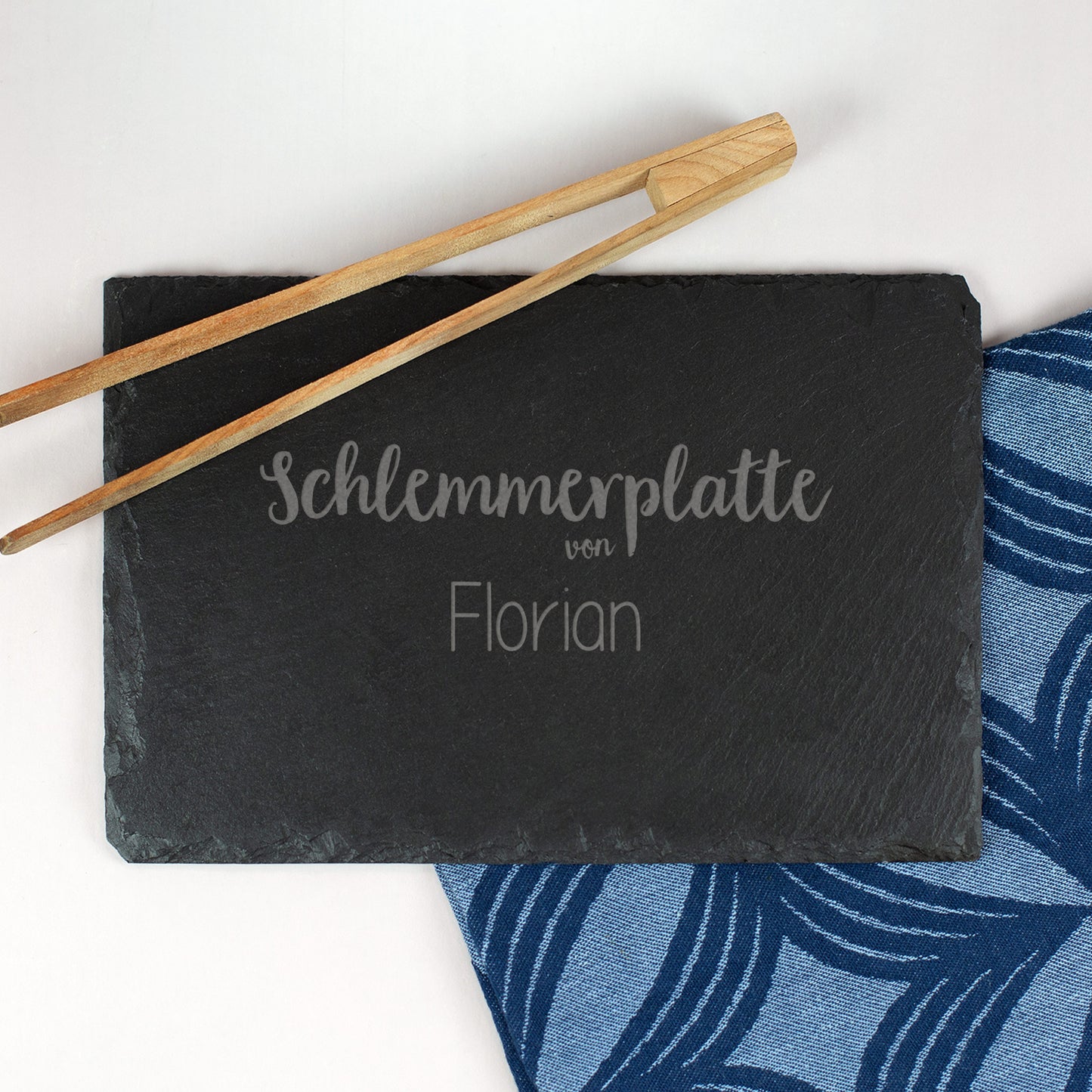 Personalisierte Schieferplatte mit Gravur „Schlemmerplatte von Florian“, daneben Holz-Grillzange und blau gemusterte Serviette.