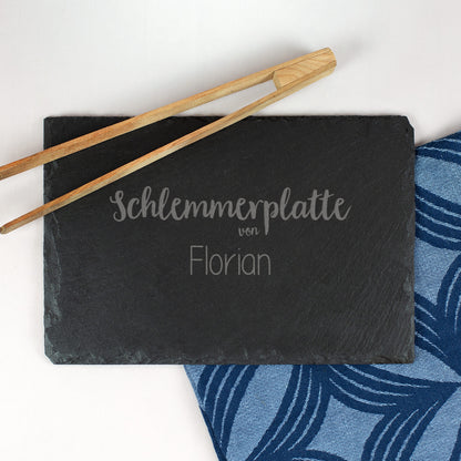 Personalisierte Schieferplatte mit Gravur „Schlemmerplatte von Florian“, daneben Holz-Grillzange und blau gemusterte Serviette.