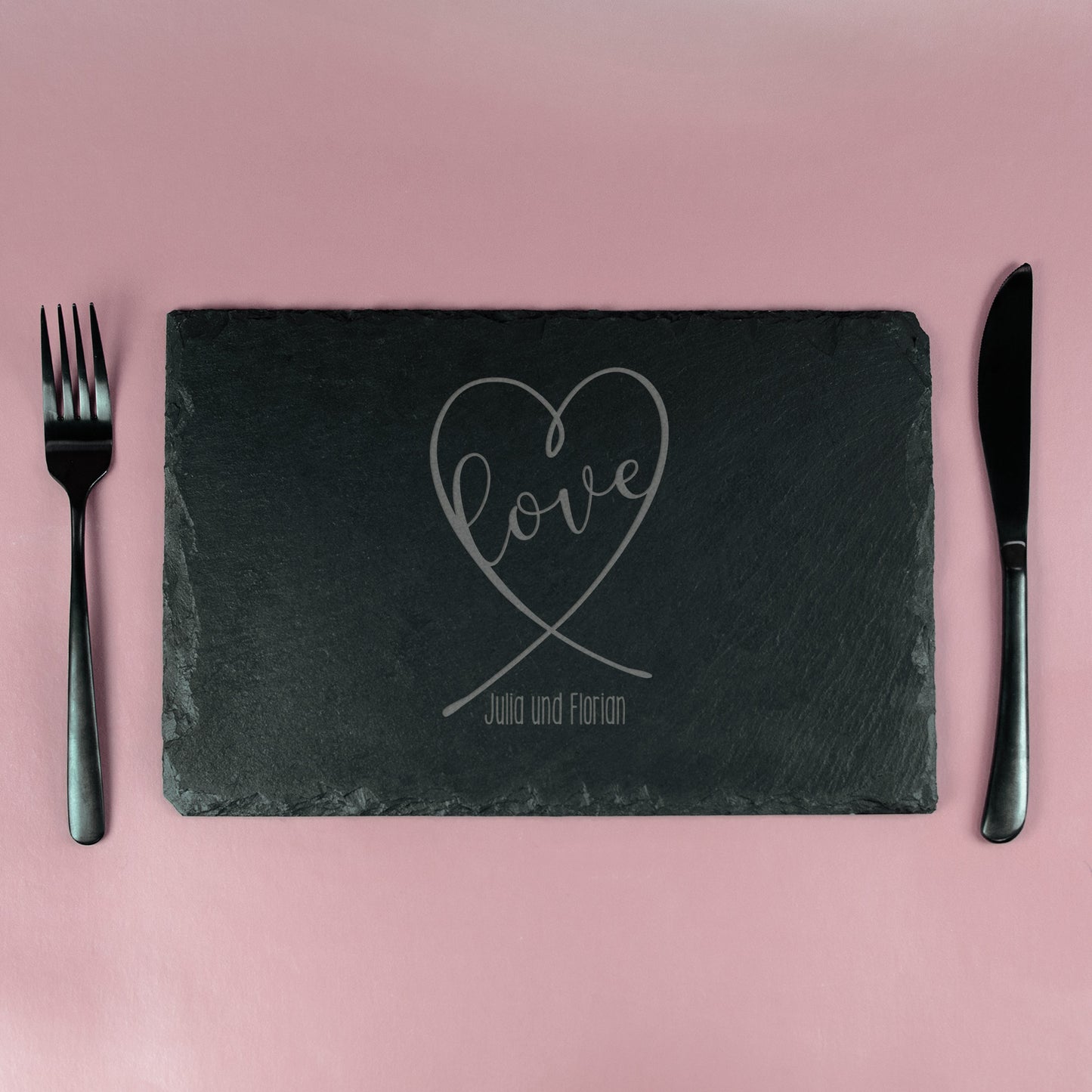 Schieferplatte mit graviertem Herz-Design und „Love“-Schriftzug, eingedeckt mit Besteck auf rosa Hintergrund