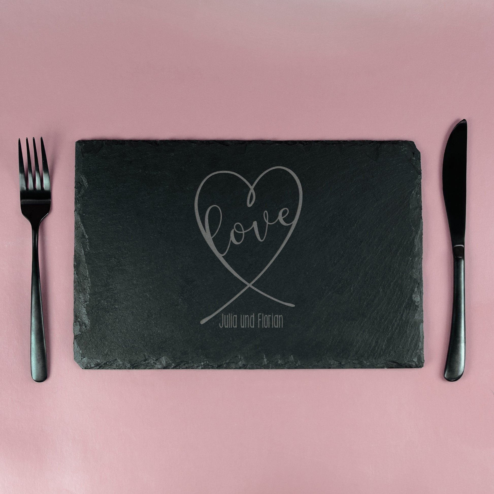 Schieferplatte mit graviertem Herz-Design und „Love“-Schriftzug, eingedeckt mit Besteck auf rosa Hintergrund