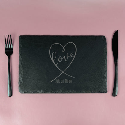 Schieferplatte mit graviertem Herz-Design und „Love“-Schriftzug, eingedeckt mit Besteck auf rosa Hintergrund