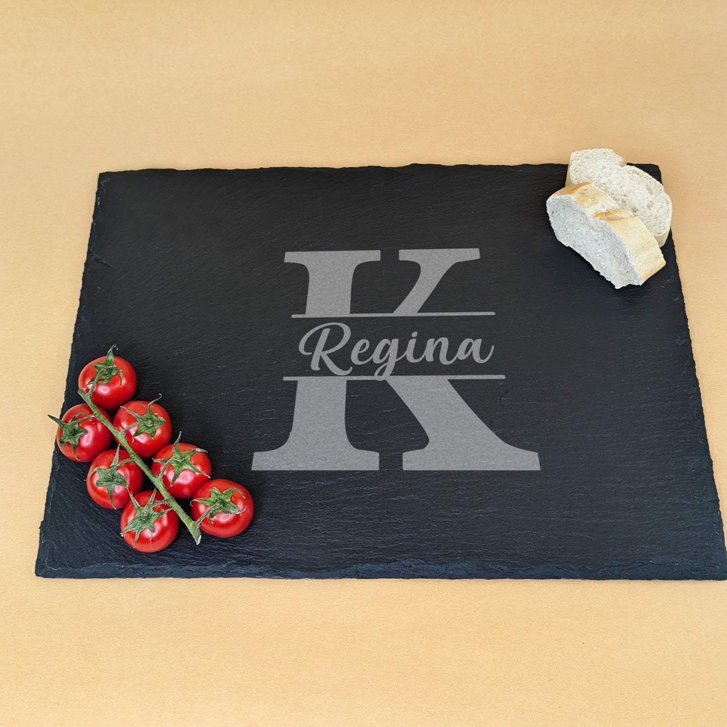Schieferplatte mit Gravur „Regina“ und Initiale „K“, dekoriert mit Tomatenrispe und Brotscheiben auf beigem Hintergrund