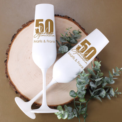 Zwei personalisierte weiße Sektgläser mit goldener Innenseite, Motiv 50 Jahre gemeinsam und Namen Marlis & Frank auf einer Holzscheibe