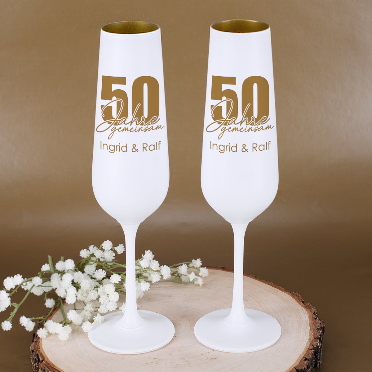 Zwei personalisierte Sektgläser zur Goldenen Hochzeit mit Motiv 50 Jahre gemeinsam und Namen Ingrid & Ralf neben weißen Blumen