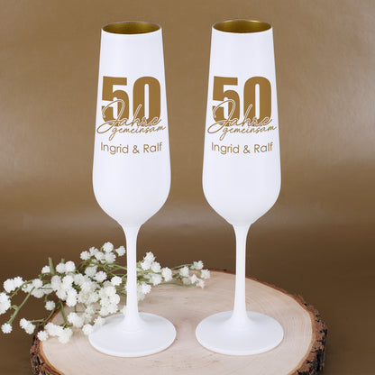 Zwei personalisierte Sektgläser zur Goldenen Hochzeit mit Motiv 50 Jahre gemeinsam und Namen Ingrid & Ralf neben weißen Blumen