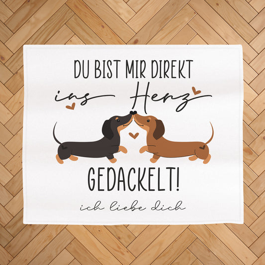 Kuscheldecke mit Dackel-Motiv auf hellem Holz-Fischgrätboden, Aufdruck „Du bist mir direkt ins Herz gedackelt!“ und Wunschtext „ich liebe dich“ unter dem Motiv
