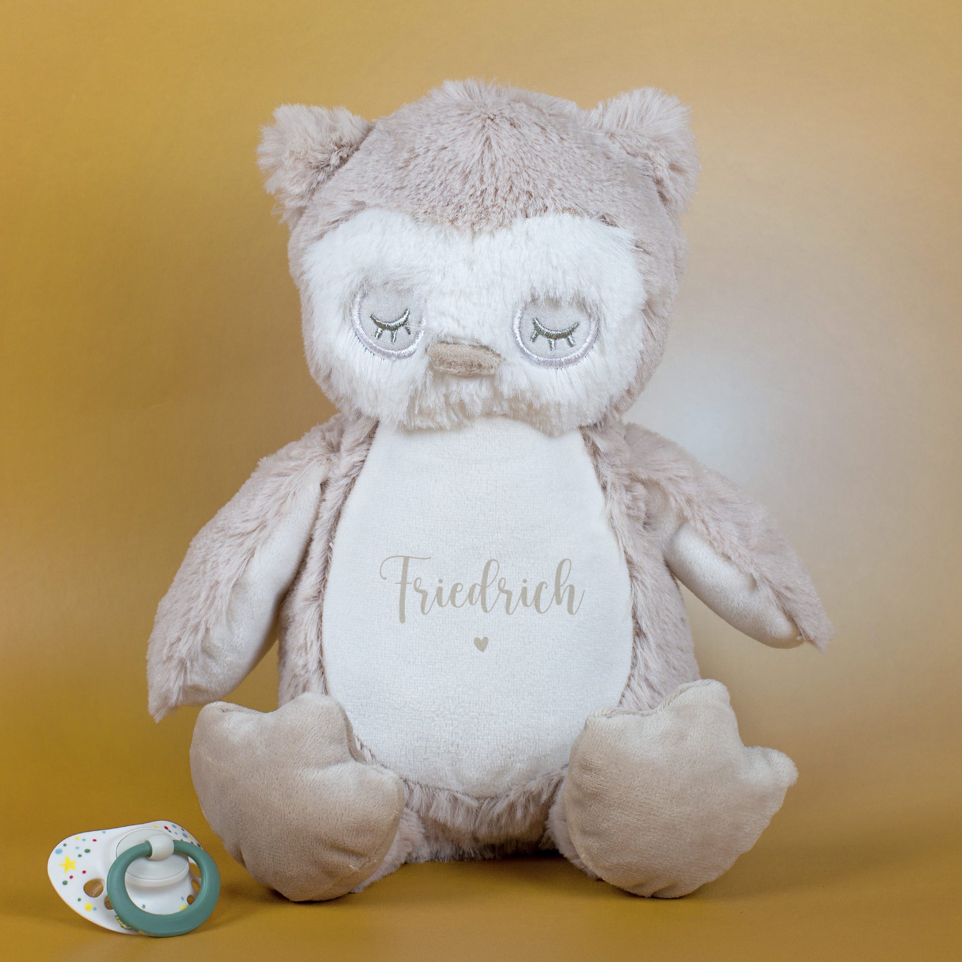 Eule mit grauer Personalisierung „Friedrich“ und Herzmotiv, sitzend vor goldenem Hintergrund mit Schnuller daneben.