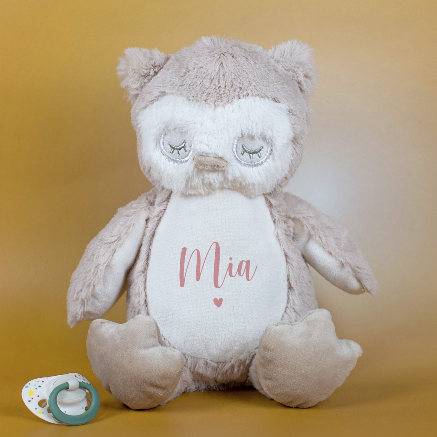 Kuscheltier-Eule mit rosa Schriftzug „Mia“ über Herzmotiv, daneben ein grüner Schnuller auf goldenem Hintergrund.