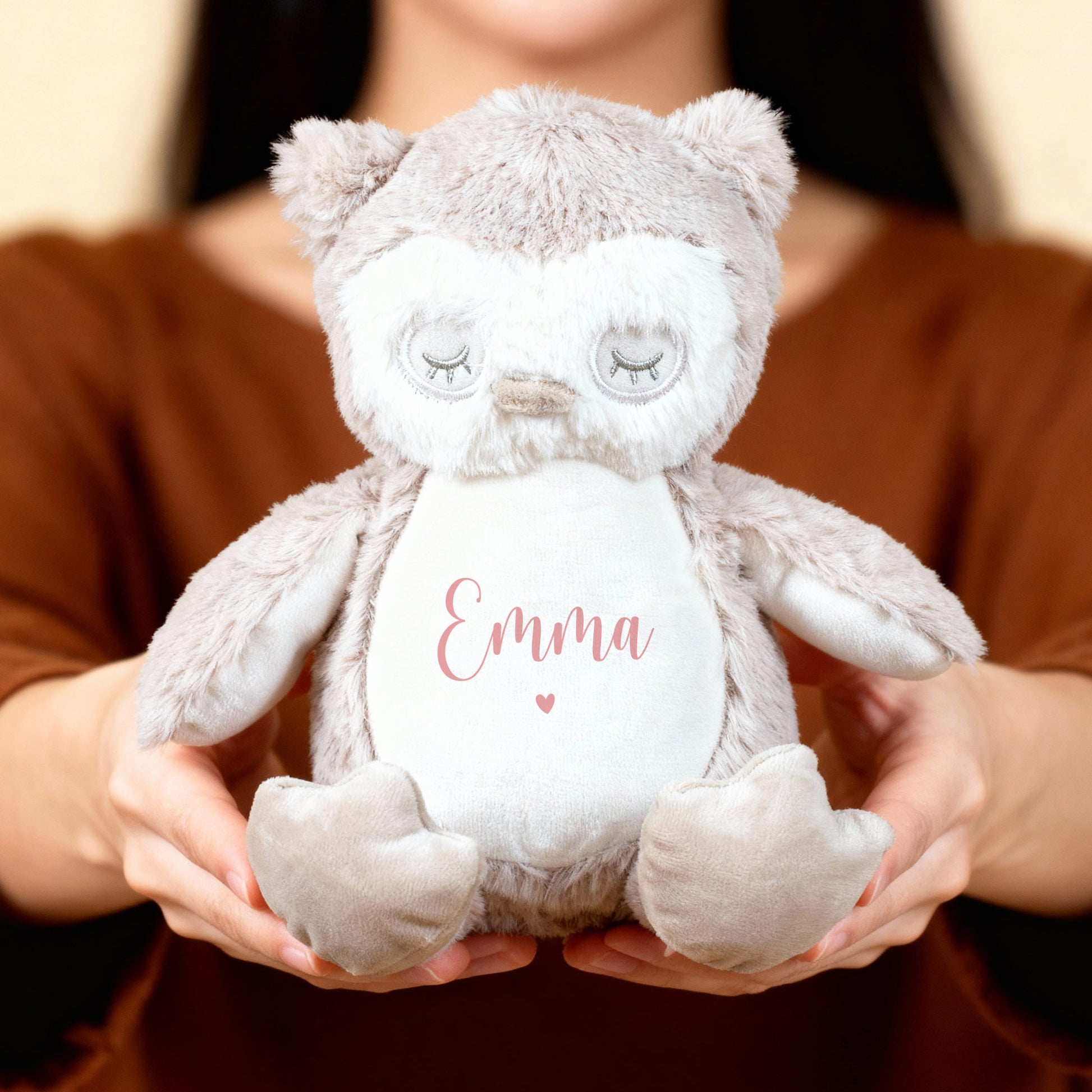 Eine Person hält eine flauschige Eule mit rosa Aufschrift „Emma“ und Herzmotiv in beiden Händen vor neutralem Hintergrund.