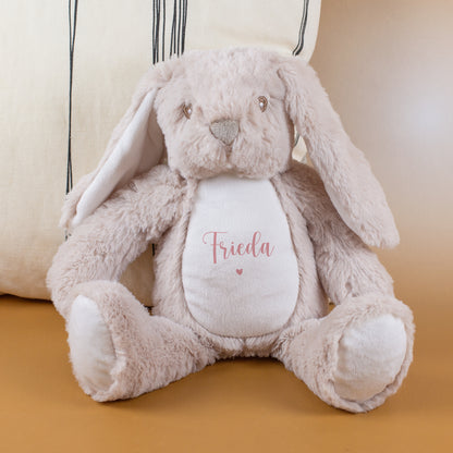 Kuscheltier-Hase mit rosa Namensaufdruck „Frieda“ und Herzmotiv sitzt auf einer beigen Unterlage vor einem hellen Kissen. Das Plüschtier ist in Beige gehalten und hat weiche, lange Ohren.