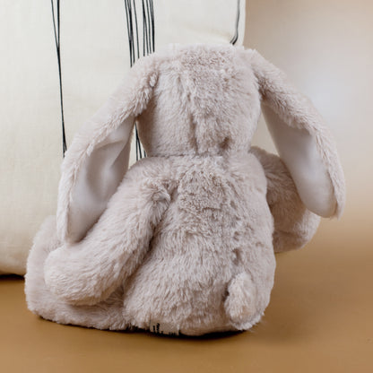 Rückansicht des flauschigen Hasen mit langen, herabhängenden Ohren und rundem Schwanz. Der Hintergrund zeigt ein helles Kissen und eine beige Fläche – Fokus auf das weiche Fell.
