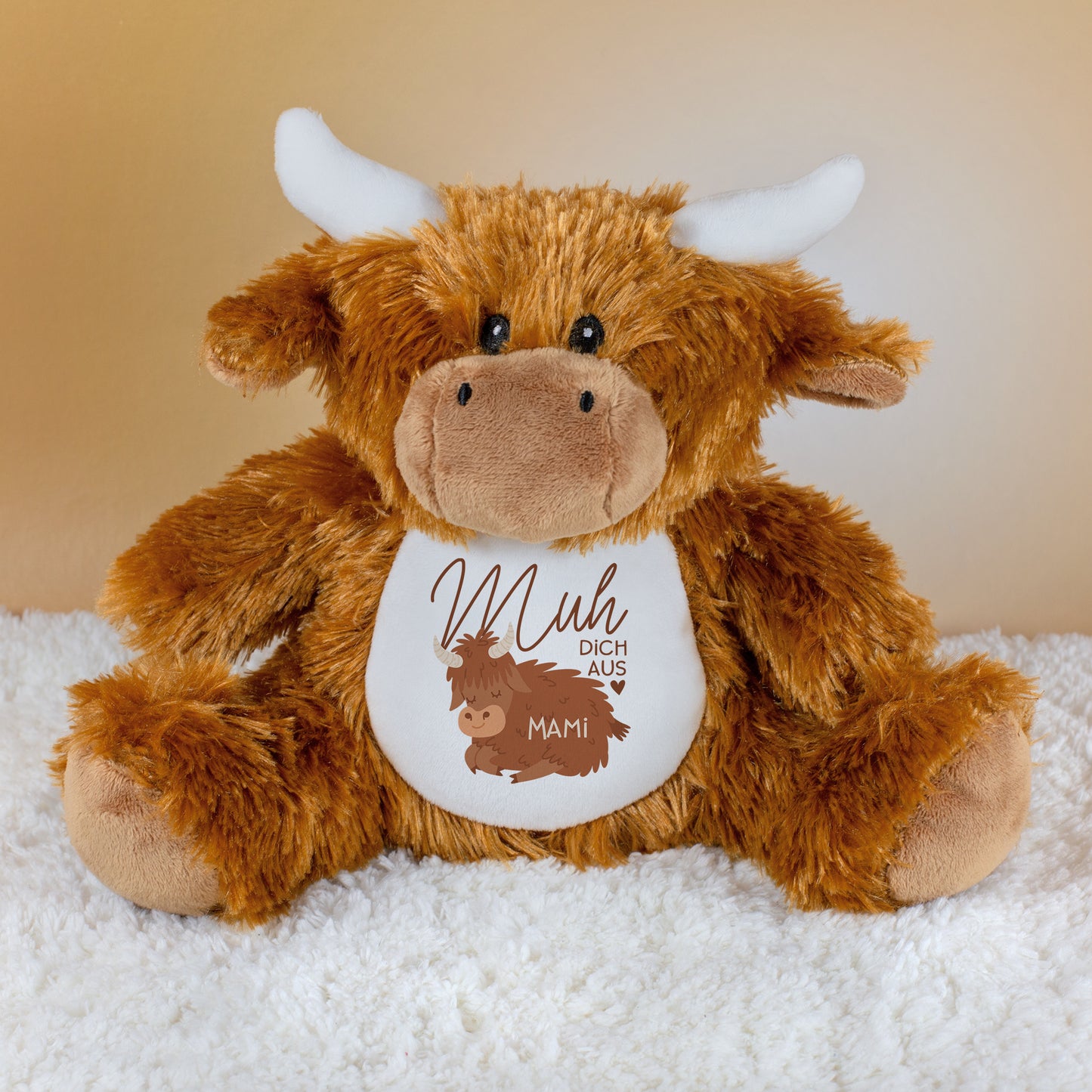 Kuscheltier-Kuh aus Plüsch mit weißem Bauch, braunem Fell und Spruch „Muh dich aus“, darunter der Name „Mami“, sitzend auf heller Unterlage.