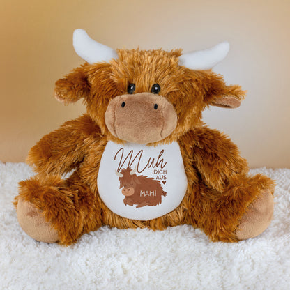 Kuscheltier-Kuh aus Plüsch mit weißem Bauch, braunem Fell und Spruch „Muh dich aus“, darunter der Name „Mami“, sitzend auf heller Unterlage.