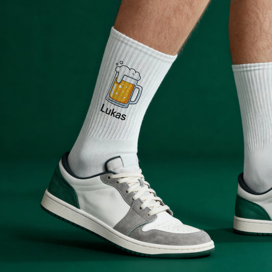 Weiße Sportsocken mit Bierkrug und Name „Lukas“ in Sneakers – Seitenansicht auf grünem Hintergrund.