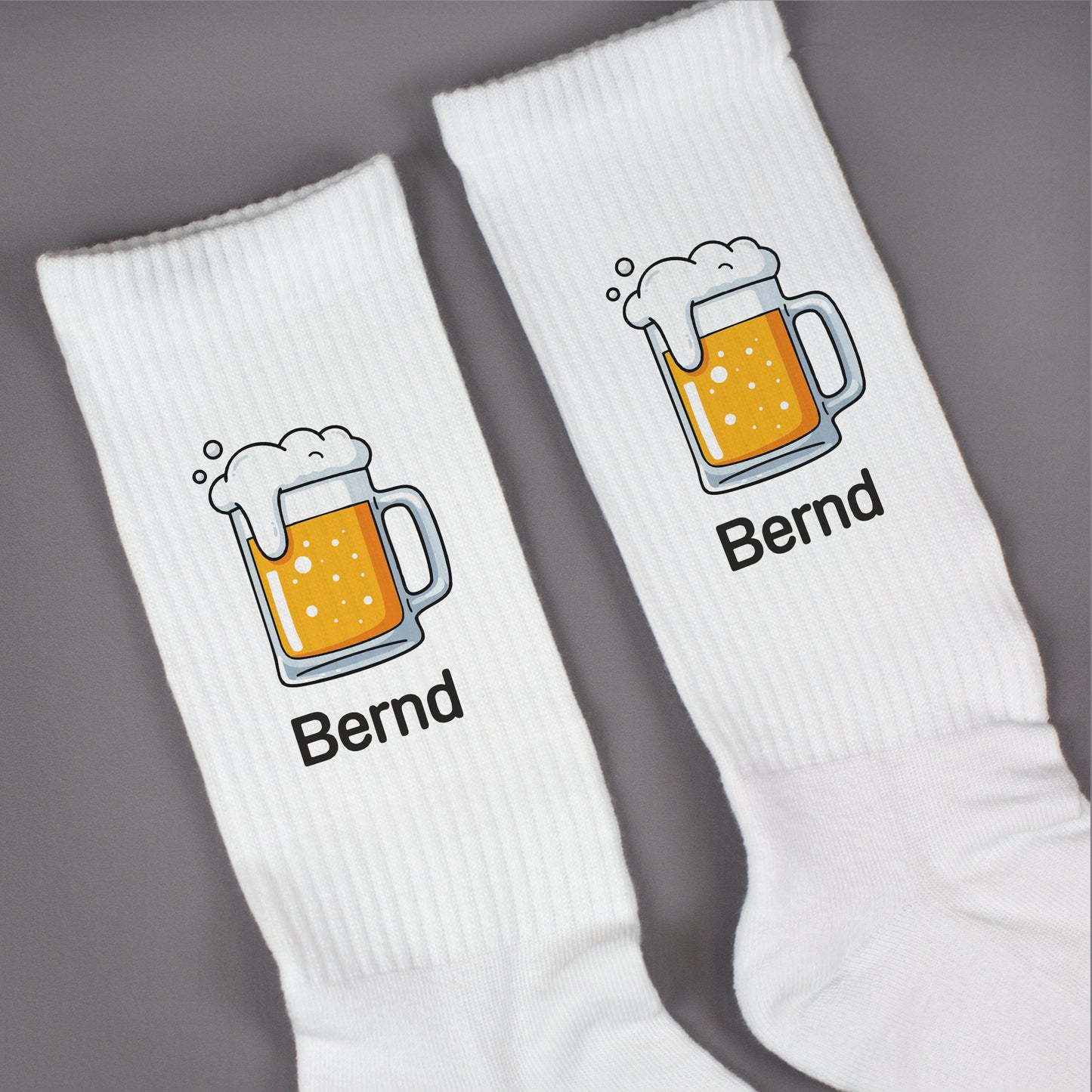 Zwei weiße Socken mit Bierkrug-Motiv und Name „Bernd“ nebeneinander auf grauer Fläche – Detailansicht des Drucks.