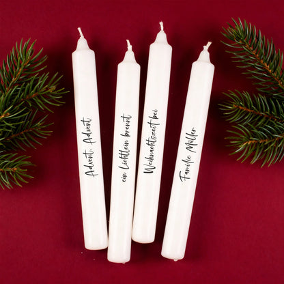 Vier personalisierte Adventskerzen mit schwarzer Schrift auf rotem Hintergrund und Tannenzweigen.