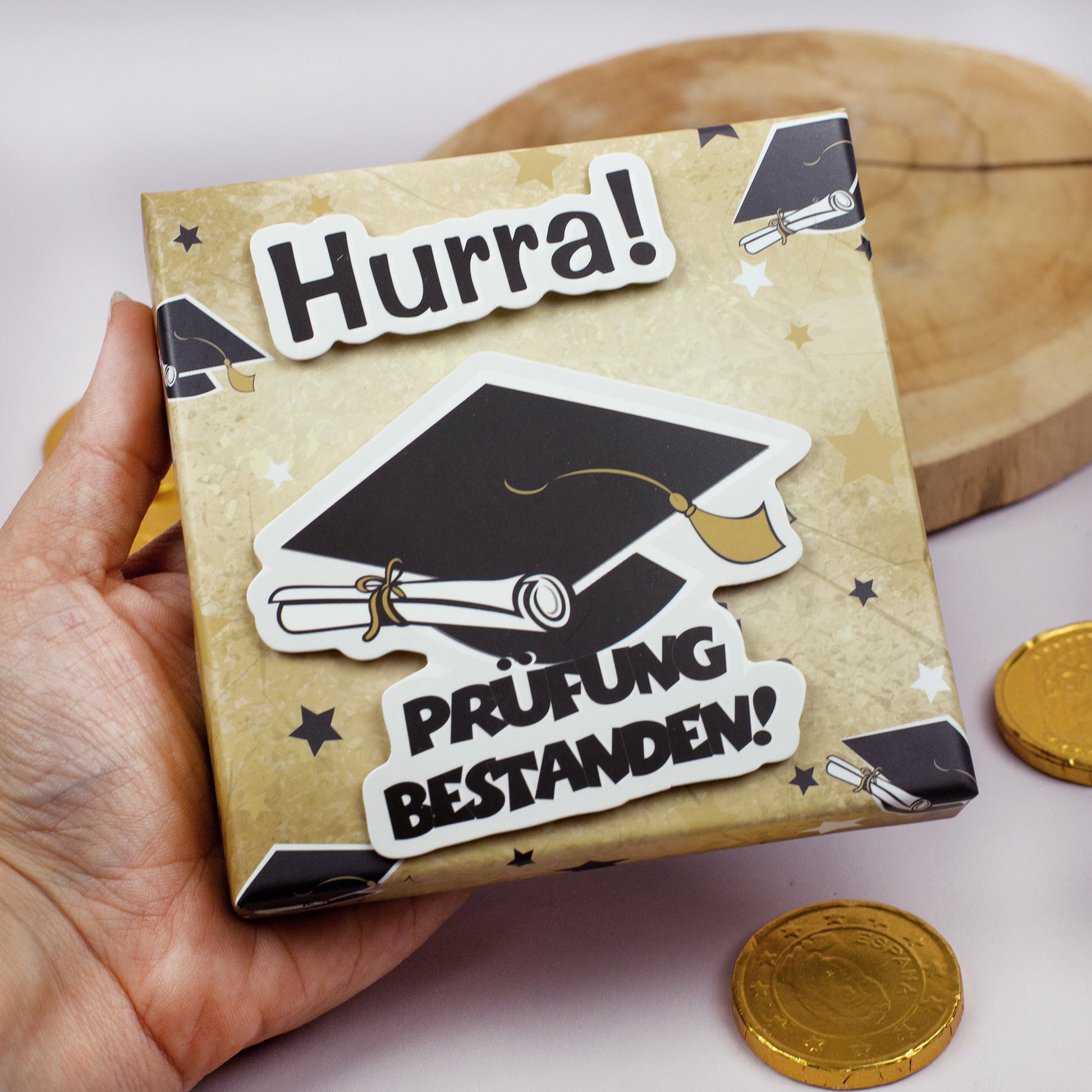 Geschenkschachtel „Hurra! Prüfung bestanden!“ mit Doktorhut und Urkunde in einer Hand gehalten
